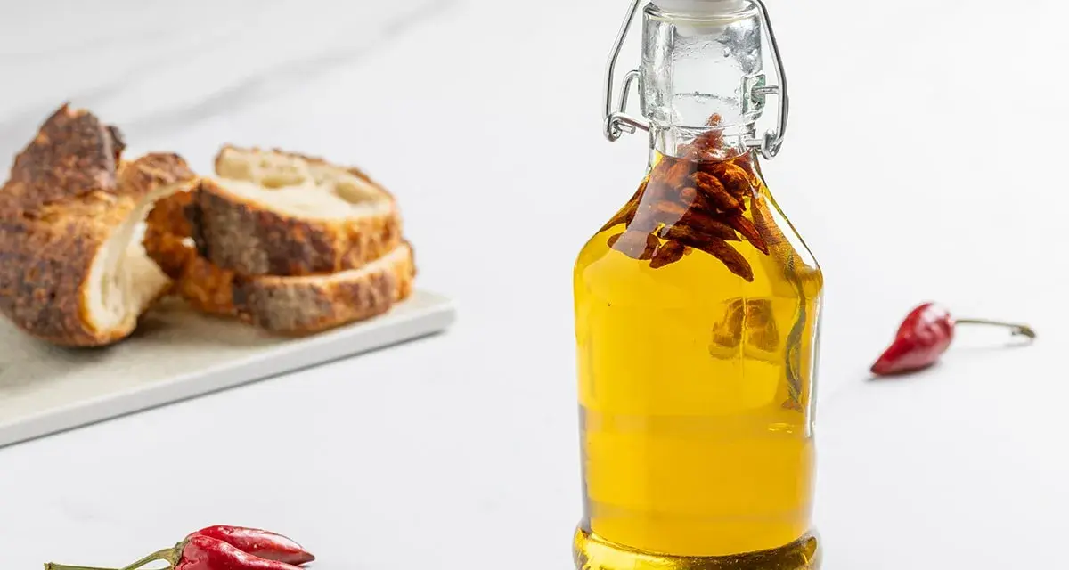 Olio al peperoncino in bottiglia, con peperoncini rossi e pane rustico a fette sullo sfondo.