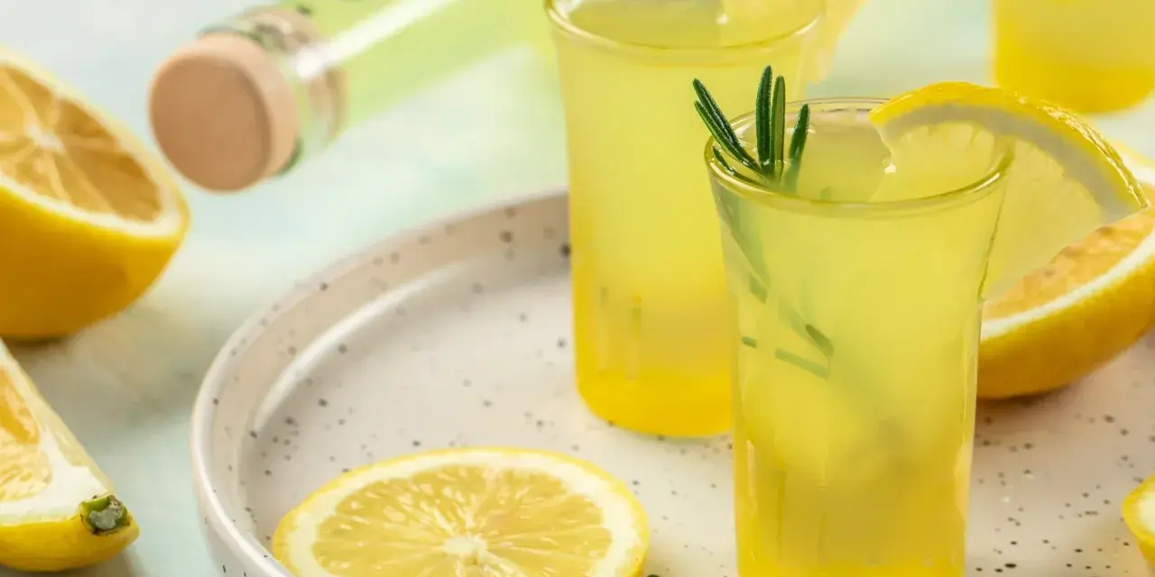 Orzeźwiający limoncello co to? Włoski likier cytrynowy w kieliszkach, ozdobiony cytryną i rozmarynem, obok butelki i świeżych cytryn.