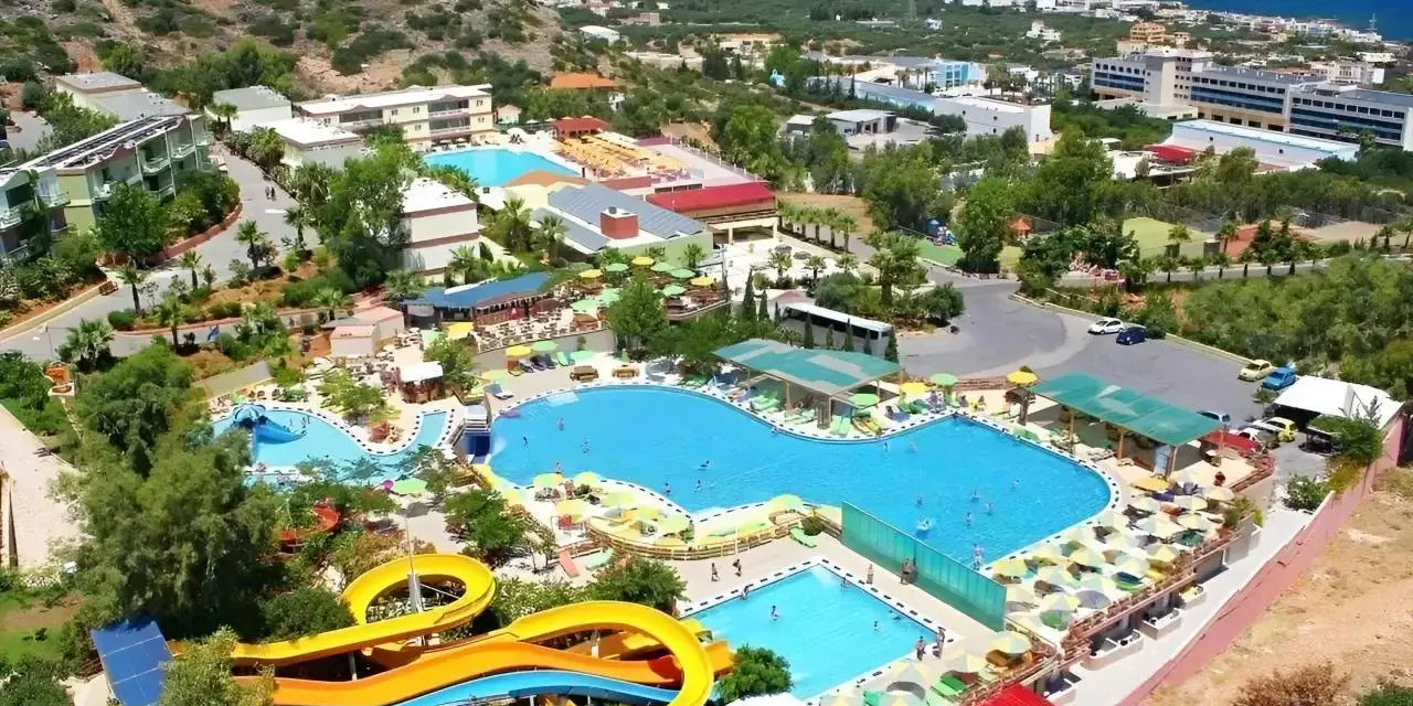 Widok z lotu ptaka na kompleks basenów z atrakcjami wodnymi i leżakami. Wiele osób korzysta z uroków słońca i wody. W tle widać budynki hotelowe i morze. Aqua Sun Village opinie potwierdzają, że to idealne miejsce na wakacje.
