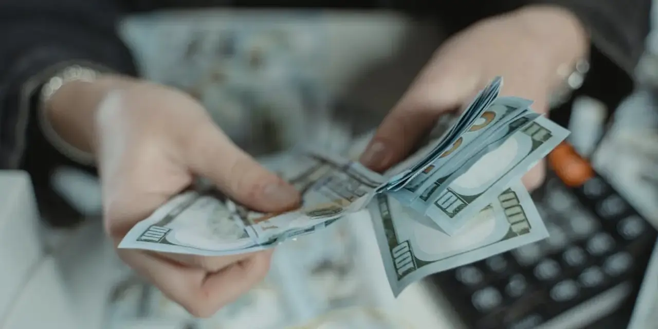 Dłonie liczą banknoty 100-dolarowe. Obok leży kalkulator, sugerując obliczanie kwoty, być może jako kara pieniężna za wyrządzoną krzywdę.