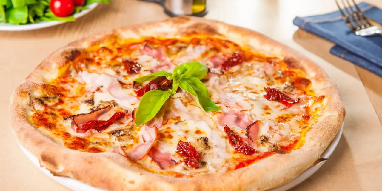 Pizza garnie de jambon, tomates séchées et champignons, parfaite pour une recette pâte à pizza rapide. Une salade fraîche est servie à côté.