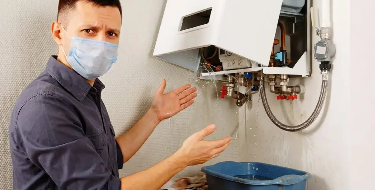 Uomo con mascherina mostra con le mani la caldaia a condensazione che perde acqua dal tubo di scarico, raccogliendola in una bacinella.