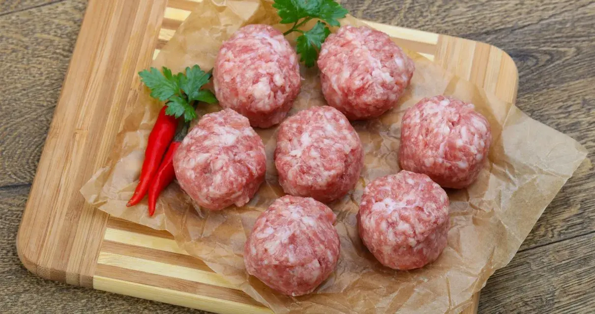 Surowe kotlety mielone keto, przyprawione papryczkami chili i natką pietruszki, leżą na papierze do pieczenia na desce.