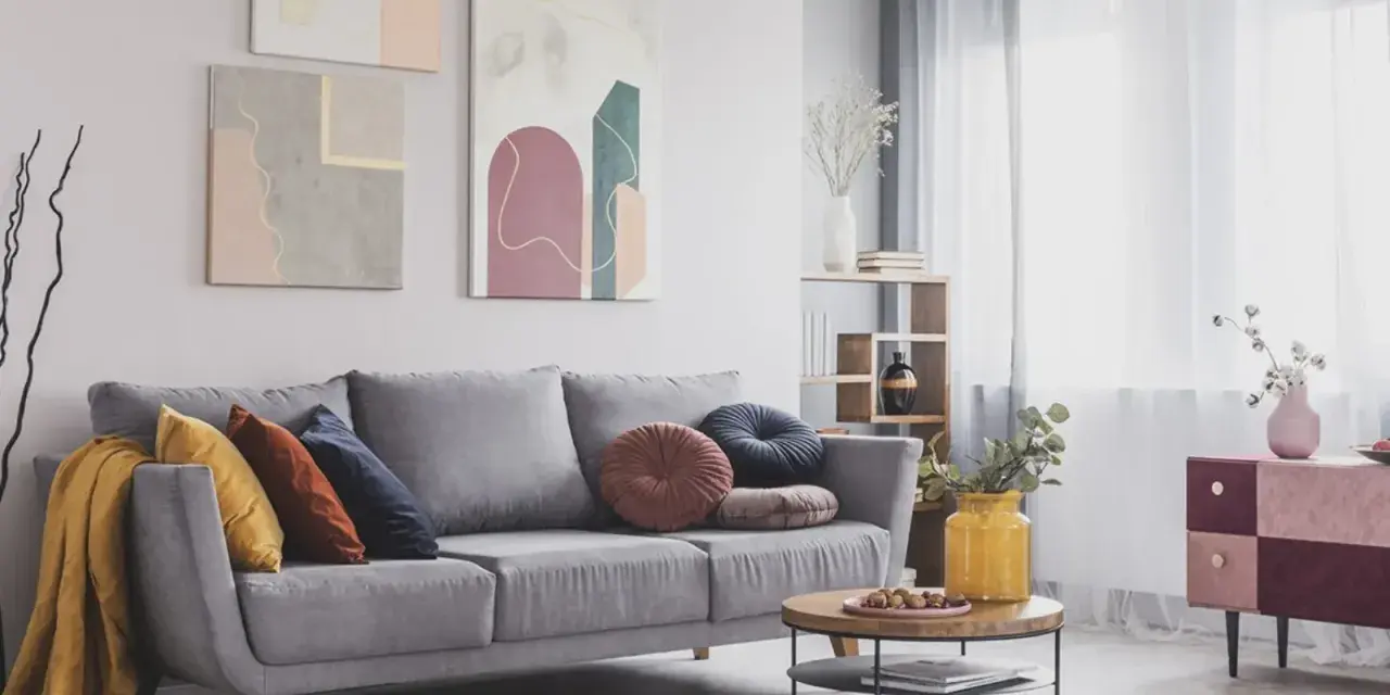 Divano grigio scuro con abbinamenti cuscini colorati e una coperta gialla. Tre quadri astratti decorano la parete.
