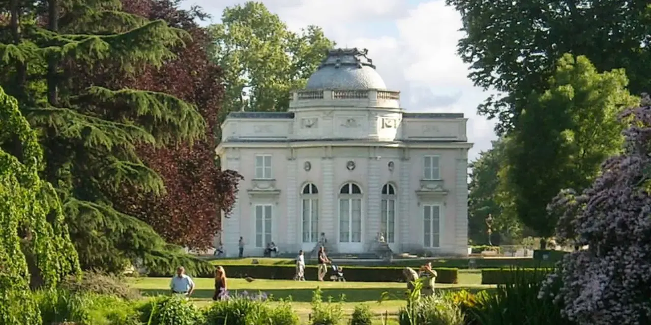 Un elegante edificio blanco con cúpula se alza en un exuberante jardin des plantes, con un estanque en primer plano y gente paseando.