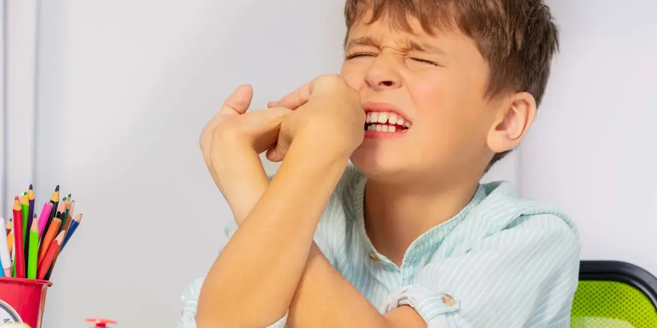 Junge mit zusammengepressten Augen und Händen vor dem Gesicht, ein Zeichen dafür, wie mit Tics bei Kindern umgehen. Buntstifte und eine Klingel liegen auf dem Tisch.