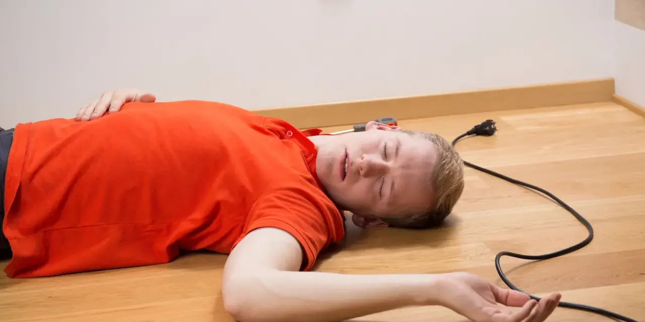 Mężczyzna w pomarańczowej koszulce leży na podłodze, obok kabel z wtyczką. Wygląda na to, że doświadczył porażenie prądem.