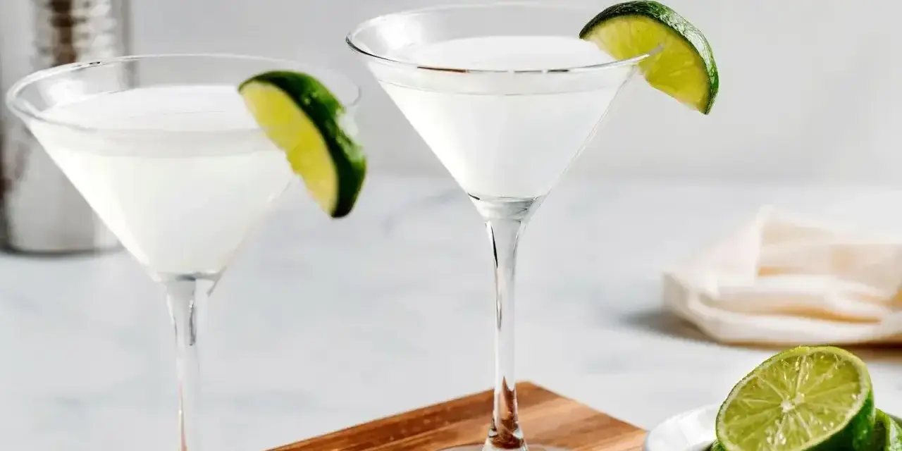 Dwa kieliszki z białym drinkiem, ozdobione plasterkami limonki. Obok miseczka z limonkami i shaker. Idealny kamikaze shot na gorący wieczór.