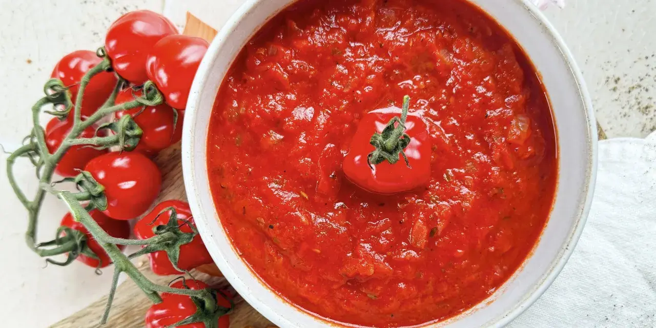 Ein Teller mit leuchtend rotem Sugo di Pomodoro, garniert mit einer einzelnen Kirschtomate. Daneben ein Bund frischer Tomaten und Zwiebeln.