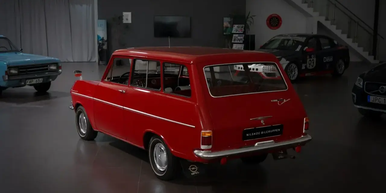 Röd veteranbil med ålder, en Opel Caravan, står i en bilhall bredvid en blå bil och en tävlingsbil.
