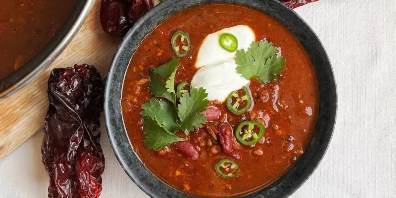 Un tazón humeante de chili con carne, adornado con crema agria, jalapeños y cilantro. Alrededor, chiles secos añaden un toque rústico.
