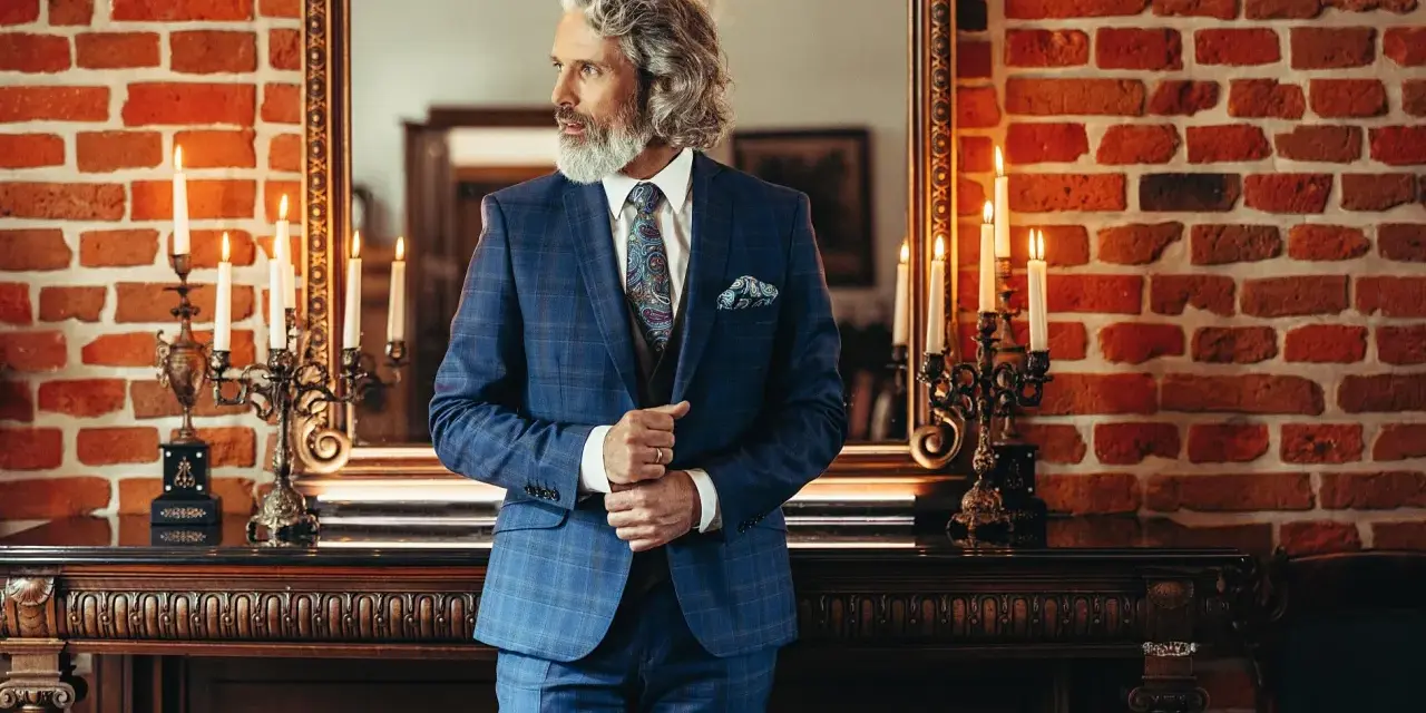 Elegancki mężczyzna w niebieskim garniturze w kratę, z wzorzystą poszetką i krawatem. Pokazuje, jak dobrać koszulę do garnituru.