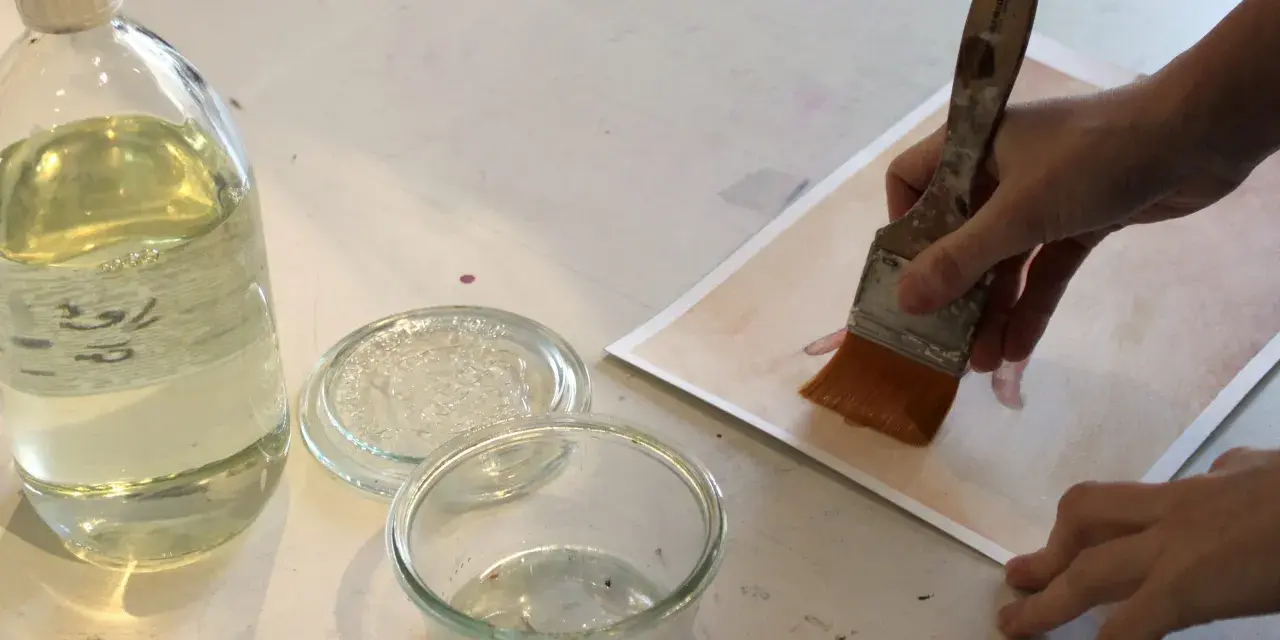 Application d'une finition acrylique sur une œuvre. Une main tient un pinceau large, prêt à recouvrir le papier d'une couche protectrice.