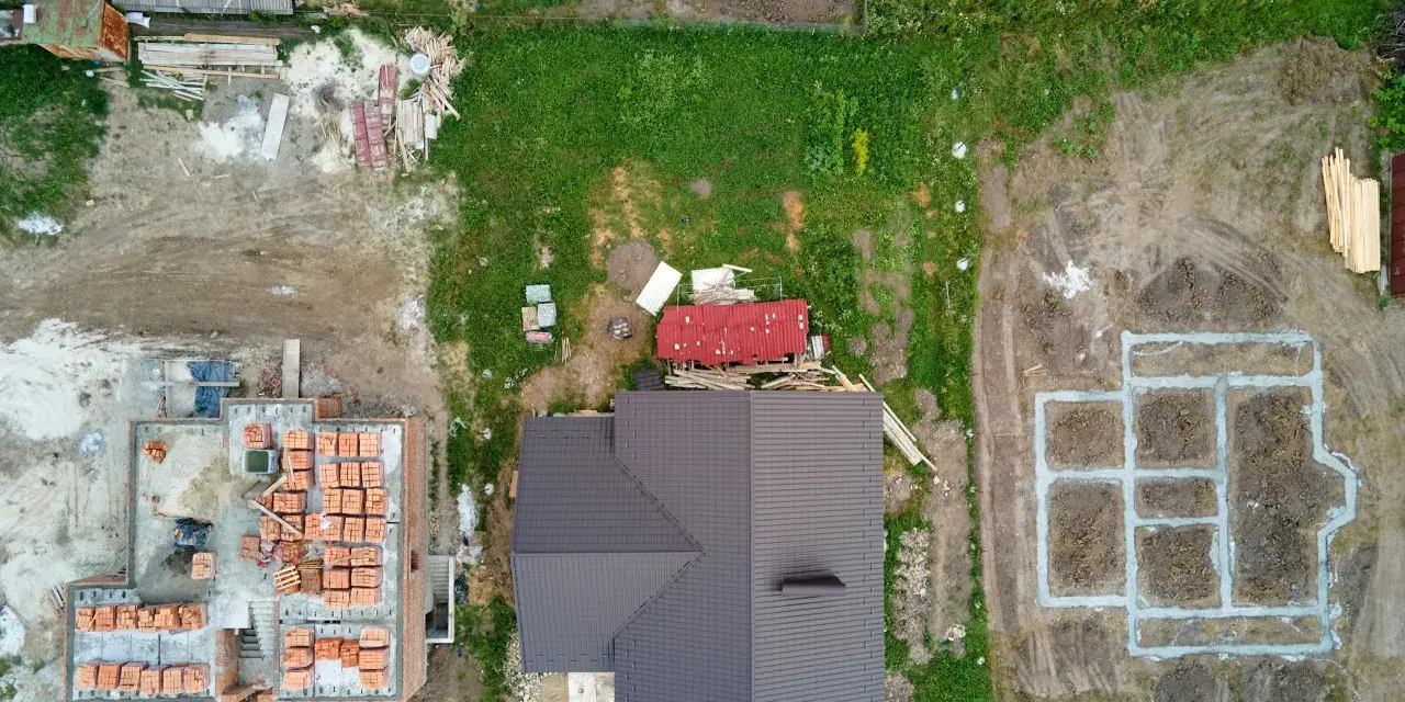Widok z lotu ptaka na plac budowy z fundamentami, częściowo zbudowanym domem i materiałami budowlanymi.