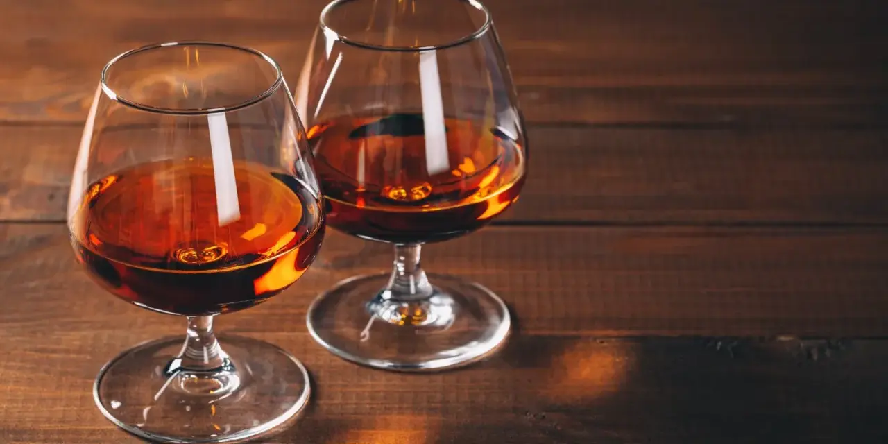Dwa kieliszki z bursztynowym płynem, przypominającym bourbon lub whiskey, stoją na drewnianym stole.