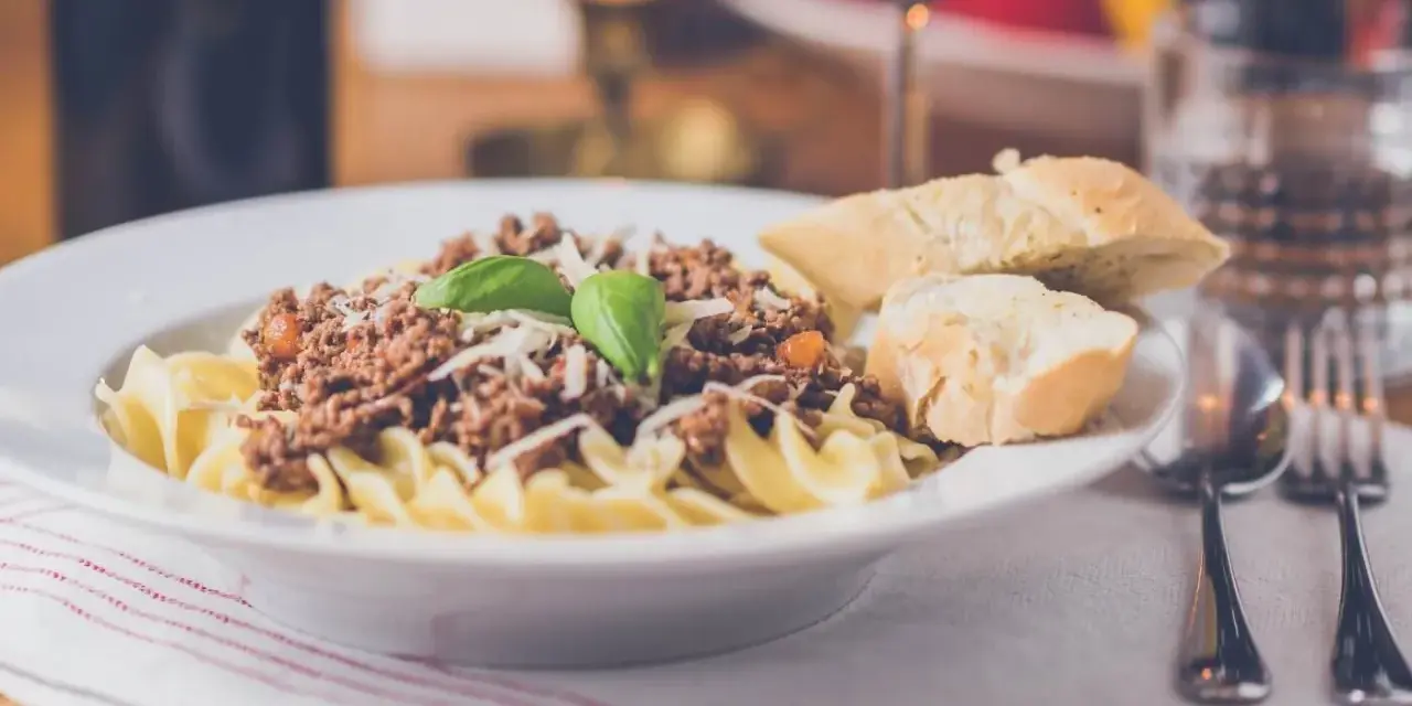 Pyszny makaron z sosem bolognese, posypany parmezanem i udekorowany bazylią. Czy to spaghetti, czy tagliatelle? Obok kromki chleba i kieliszek czerwonego wina.