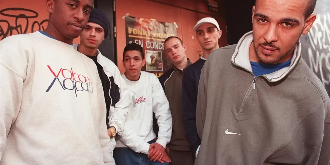 Un groupe de rap français des années 90/2000 pose devant un mur tagué. Ils portent des sweats et des jeans, typiques de l'époque.