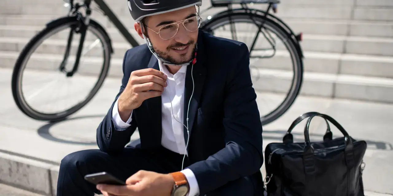 Mężczyzna w garniturze i kasku siedzi na schodach z rowerem w tle, słuchając muzyki przez słuchawki i przeglądając telefon.