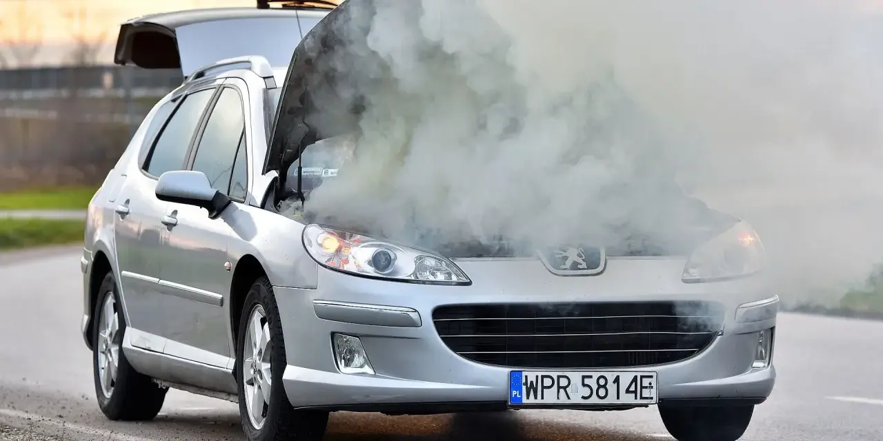 Srebrny samochód Peugeot z otwartą maską, z której wydobywa się gęsty dym.