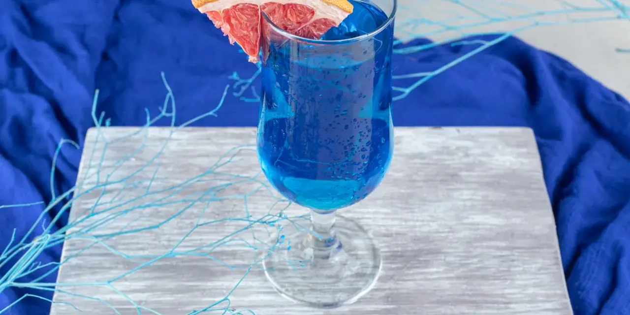 Przepis na Blue Lagoon: Błękitny drink z plasterkiem grejpfruta, ozdobiony niebieskimi gałązkami, na tle granatowej tkaniny.