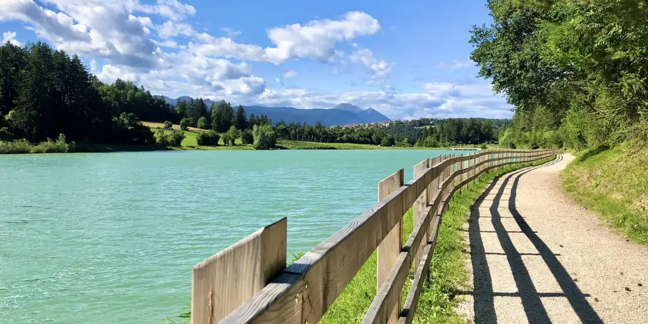Sentiero sterrato con staccionata in legno lungo il lago di Tavon, circondato da alberi e montagne. Cielo azzurro con nuvole bianche.