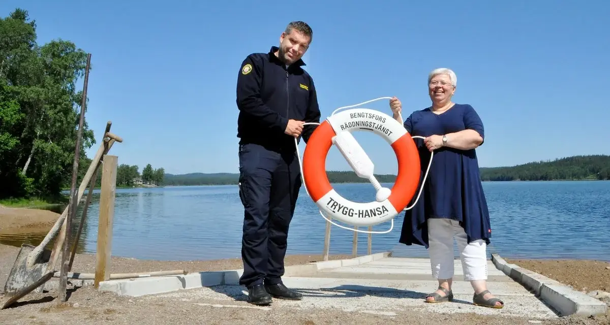 Två personer håller i en räddningsboj vid en sjö. En man i uniform och en kvinna ler. Trygg-Hansa vid Nörybyns camping.
