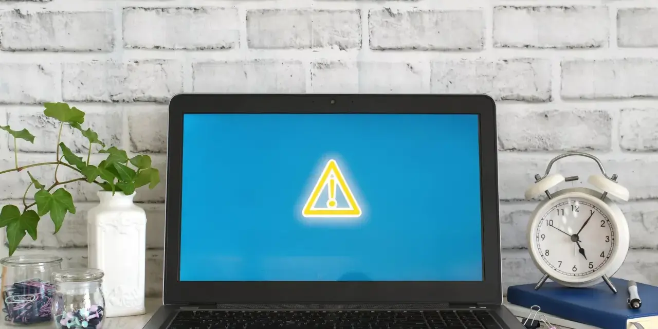 Laptop z symbolem ostrzeżenia na ekranie, obok budzik i rośliny.