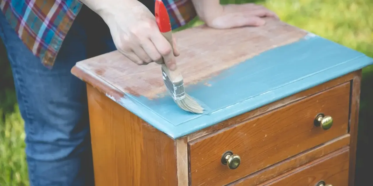 Persona pinta con brocha azul una mesita de noche de madera, transformándola en una de las mesitas de noche pintadas a la tiza.