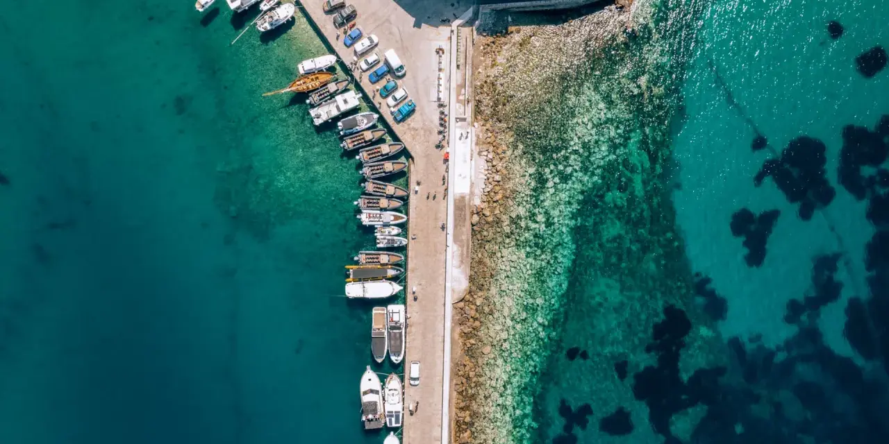 Widok z lotu ptaka na port z licznymi łodziami i jachtami cumującymi przy nabrzeżu. Woda jest krystalicznie czysta, a na dnie widać kamienie i ciemne plamy wodorostów.