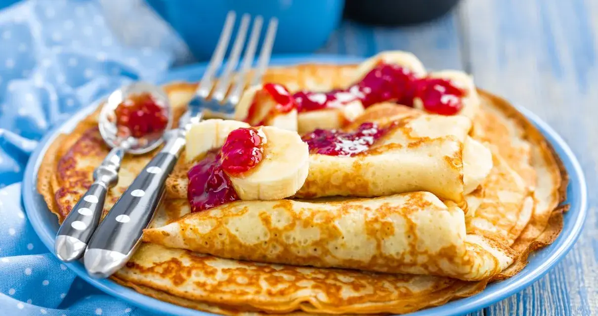 Stos idealnie przygotowanego ciasta na naleśniki, polanego dżemem i kawałkami banana. Obok leżą sztućce.