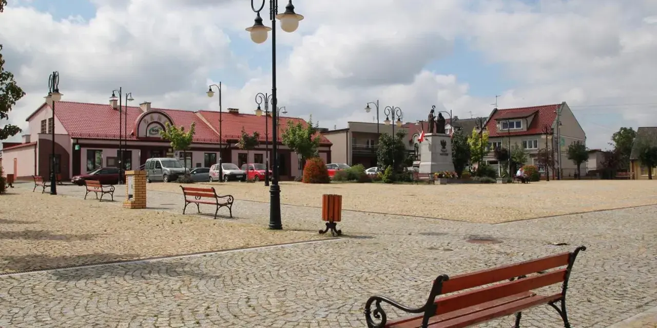 Rynek w Ślesinie z pomnikiem, ławkami i budynkami. Atrakcje tego miejsca zachęcają do odpoczynku.