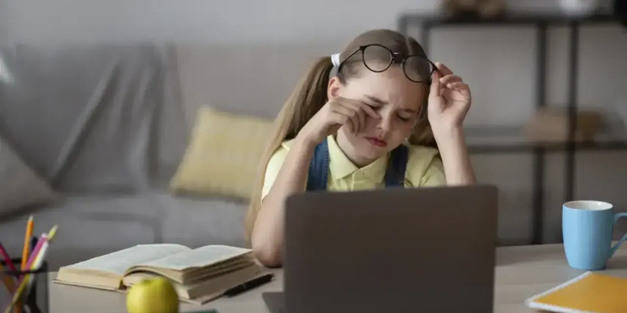 Zmęczona dziewczynka z okularami na czole pociera oczy, ucząc się przy laptopie. Jej indywidualna ścieżka nauczania jest trudna.