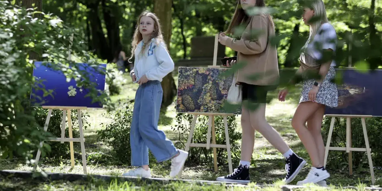 Trzy dziewczyny spacerują wśród obrazów na sztalugach. Jedna z nich, w niebieskich jeansach, przypomina młodszą wersję Tomasza Kłosowskiego.