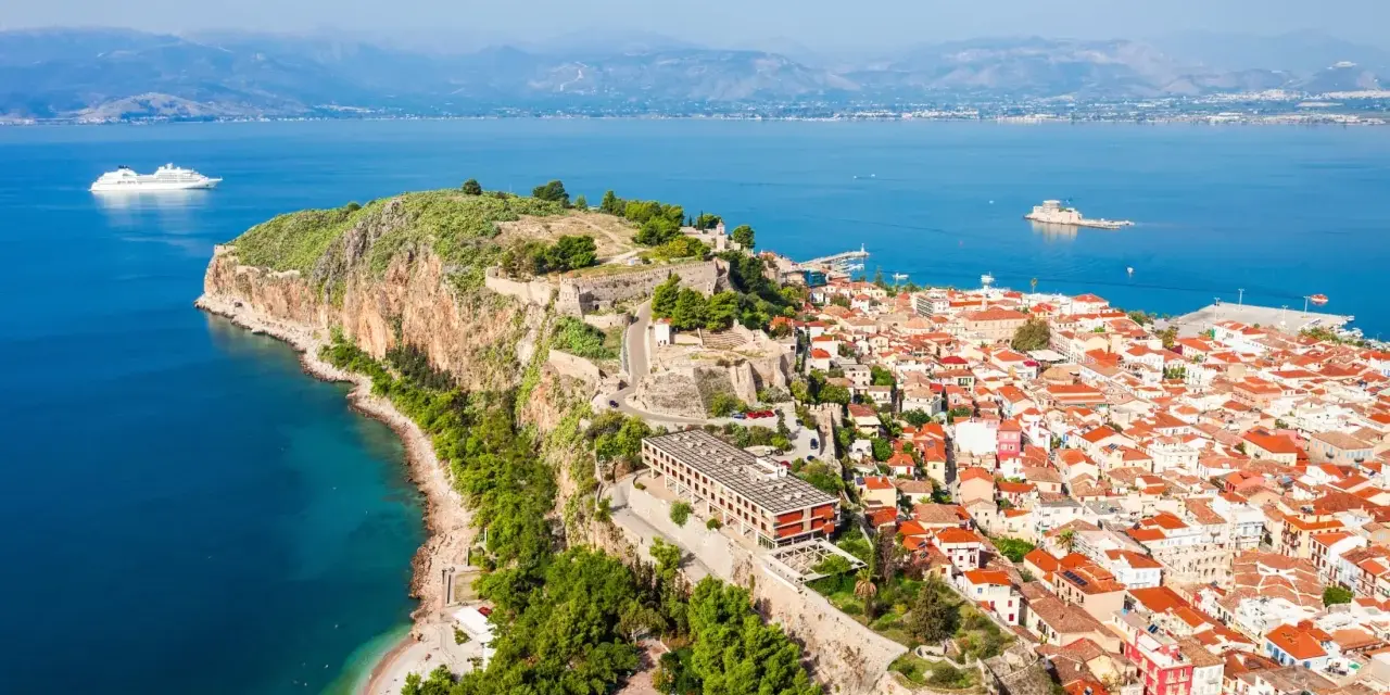 Widok na Nafplio w Grecji: miasto z czerwonymi dachami, twierdza na skale i biały statek wycieczkowy na lazurowym morzu.