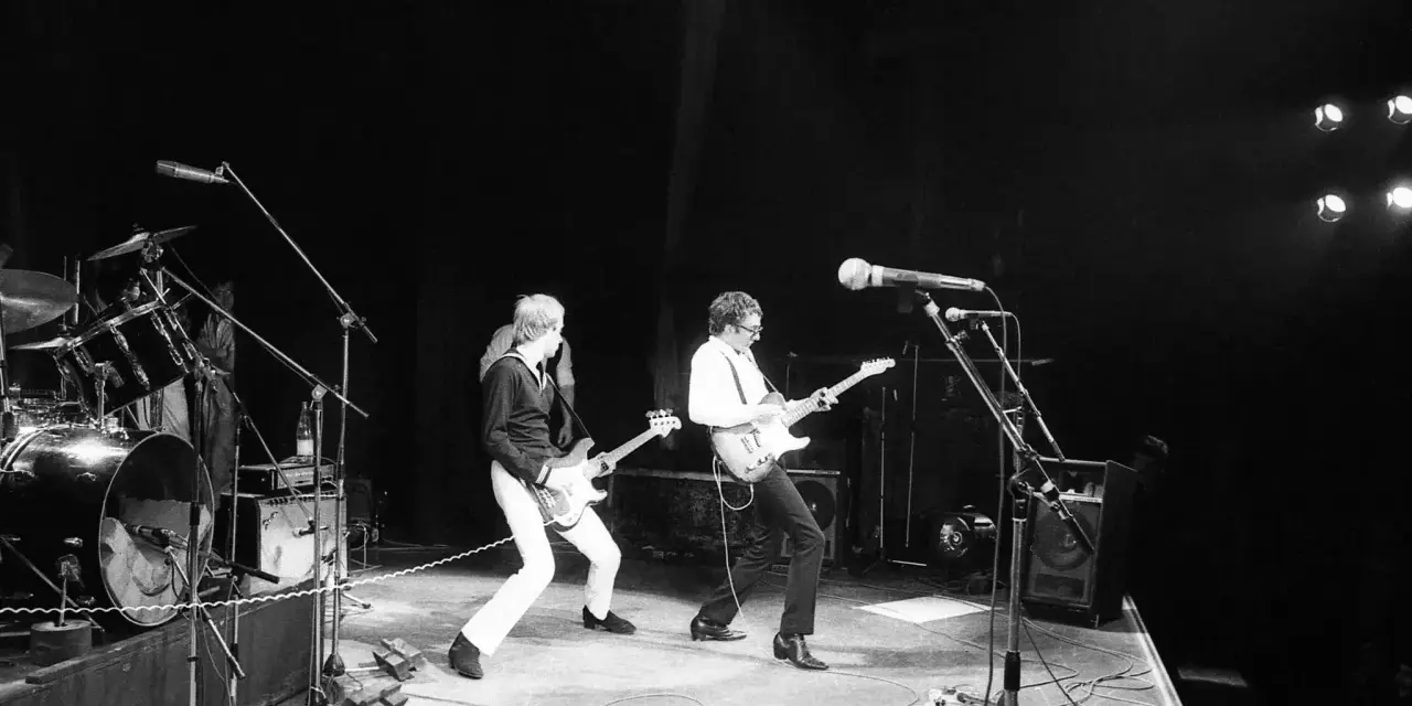 Deux guitaristes d'un groupe français rock jouent sur scène, éclairés par des projecteurs.