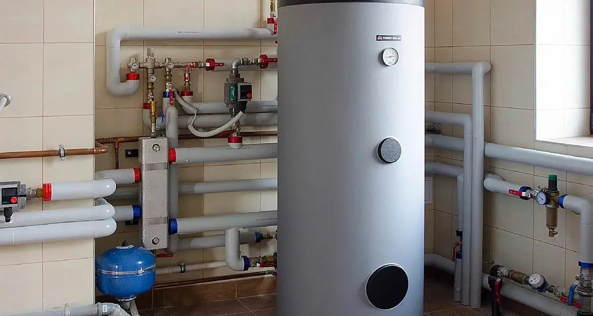 Duży, szary bojler stojący obok instalacji hydraulicznej z rurami i manometrami.