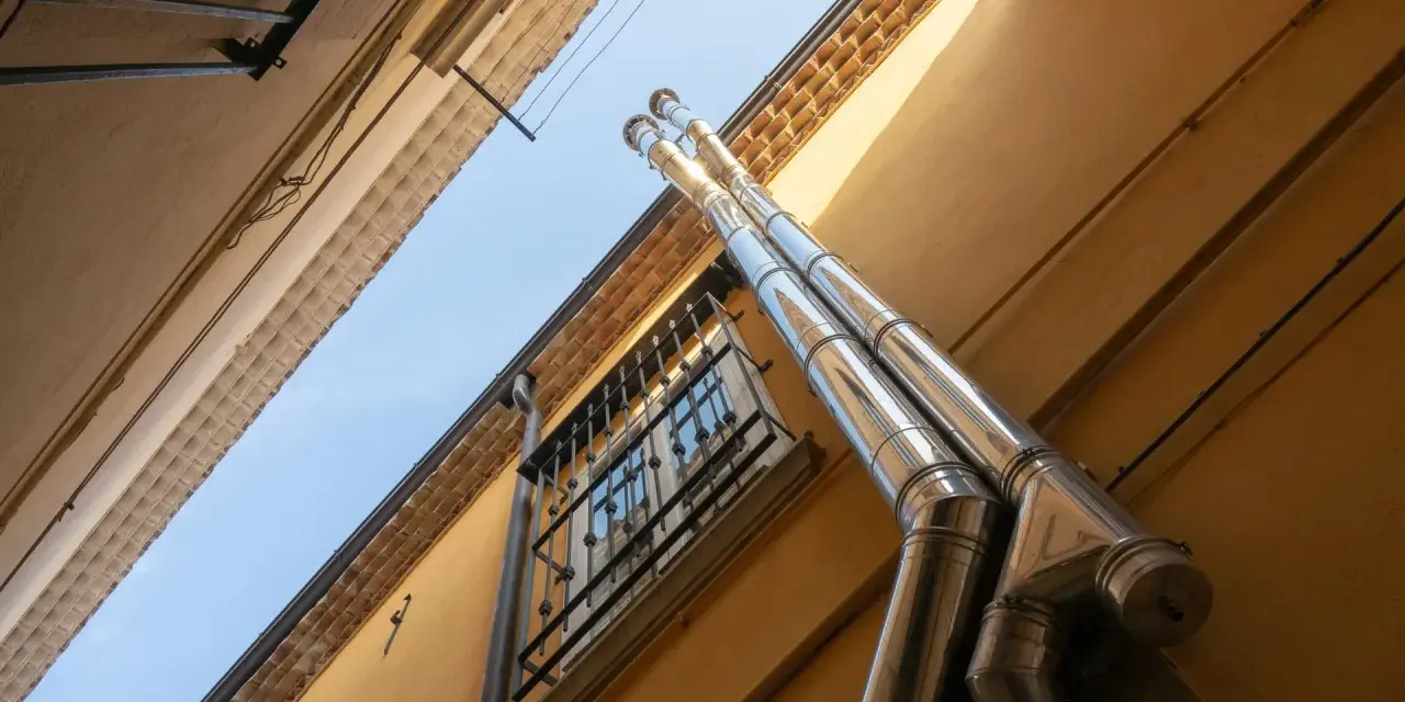 Dos tubos de salida humos estufa pellets brillan bajo el sol, ascendiendo por la pared amarilla de un edificio.