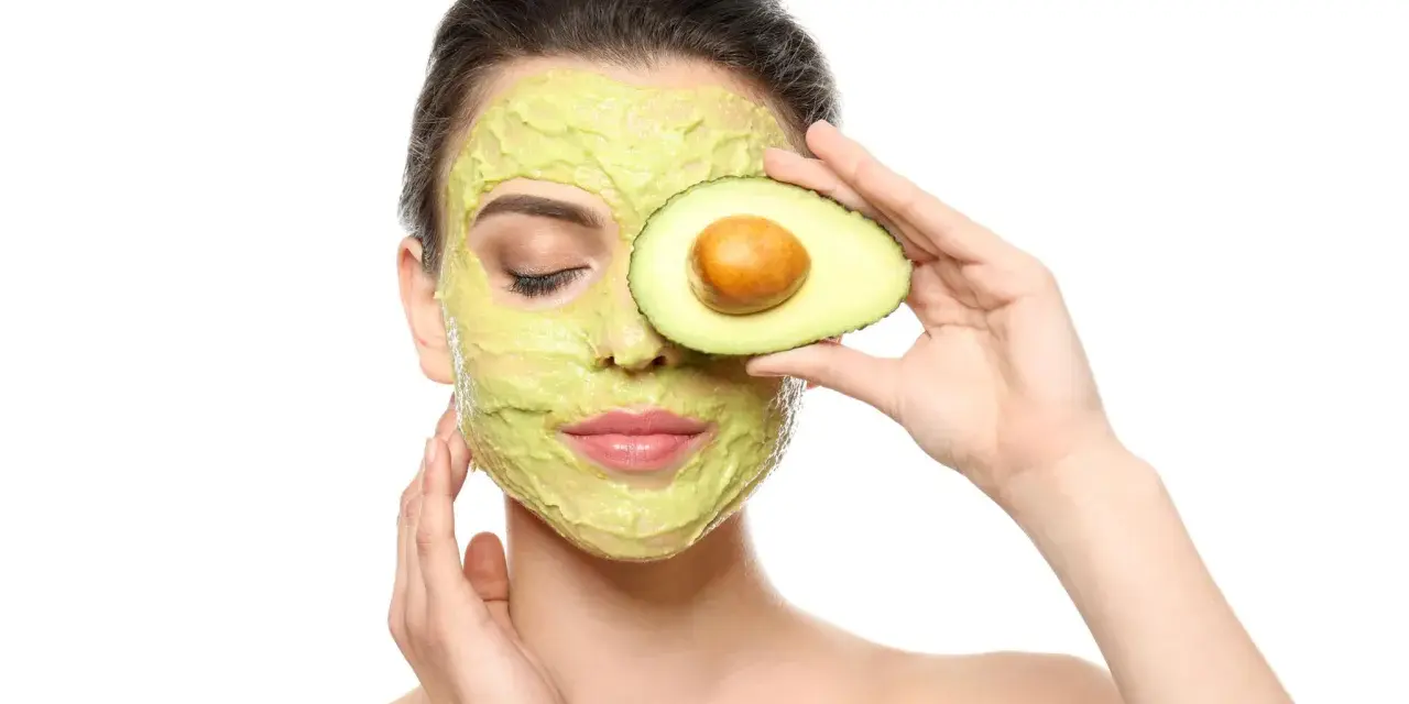 Frau mit Avocado-Gesichtsmaske und Avocado-Scheibe vor dem Auge. Die Avocado-Wirkung auf die Haut ist bekannt für ihre pflegenden Eigenschaften.