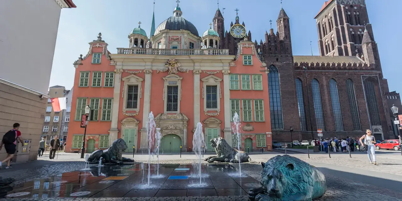 Fontanna Czterech Kwartałów w Gdańsku z lwami i wodnymi strumieniami przed różową kamienicą i bazyliką.