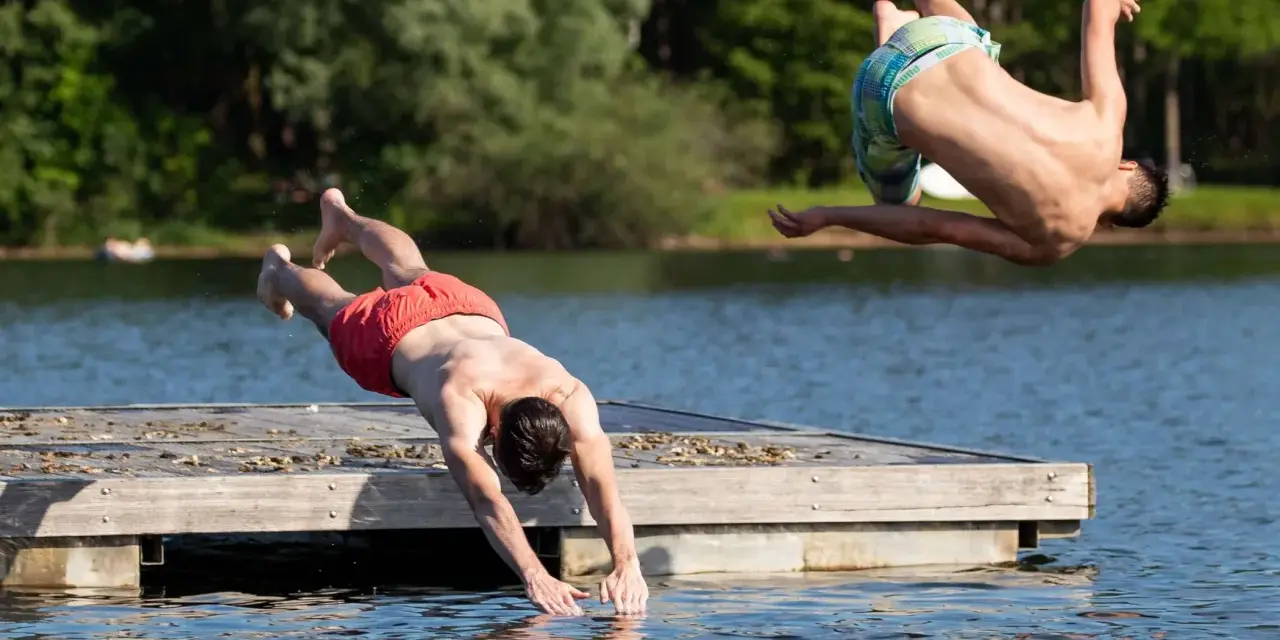 Zwei Männer springen vom Steg in den Sieglarer See. Einer macht einen Salto, der andere taucht gerade ins Wasser.