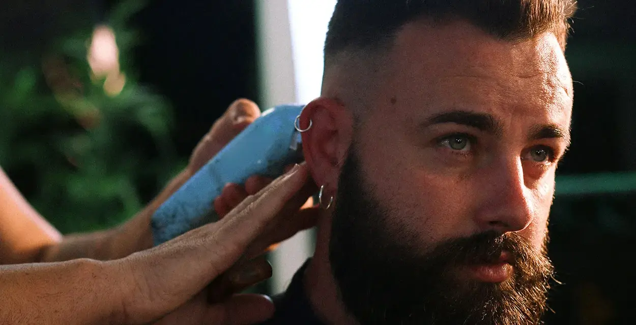 Un hombre con barba y aros en las orejas recibe un corte de pelo moderno, anticipando las tendencias de corte de pelo hombre 2026.