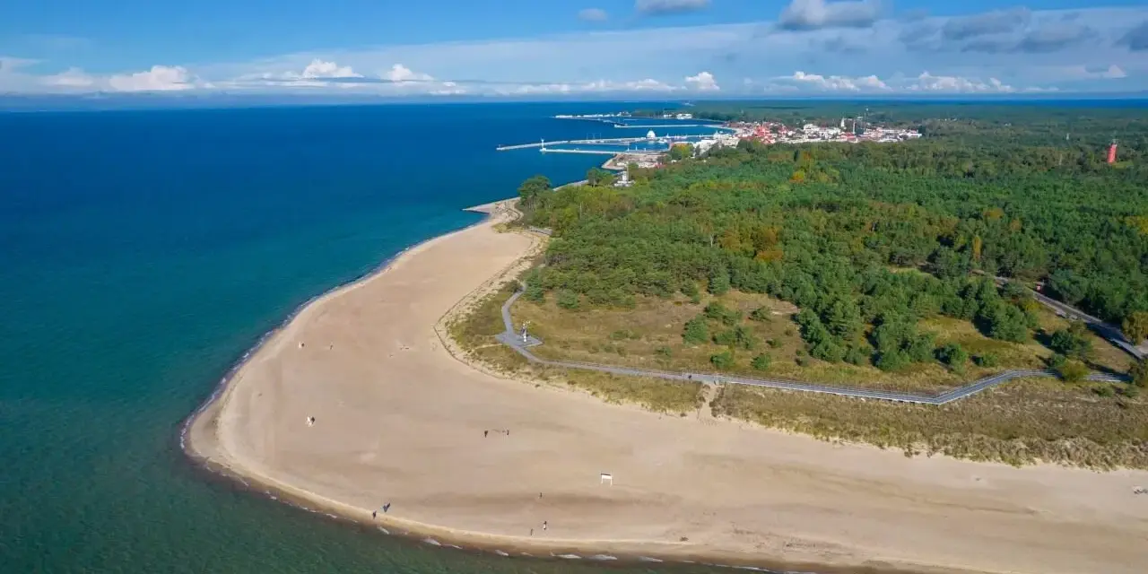 Piękna plaża i las sosnowy nad morzem. W oddali widać miasteczko i port. To właśnie tutaj jest Hel.