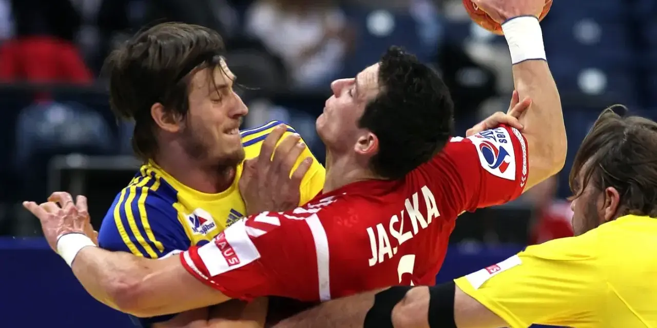 Zacięta walka w meczu polska szwecja piłka ręczna. Zawodnicy walczą o piłkę, jeden w czerwonej koszulce z napisem Jaszka, drugi w żółtej.