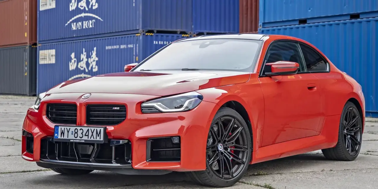 Pomarańczowe BMW M2 z czarnymi felgami, na tle niebieskich kontenerów. Sprawdź, jaka jest aktualna bmw m2 cena.