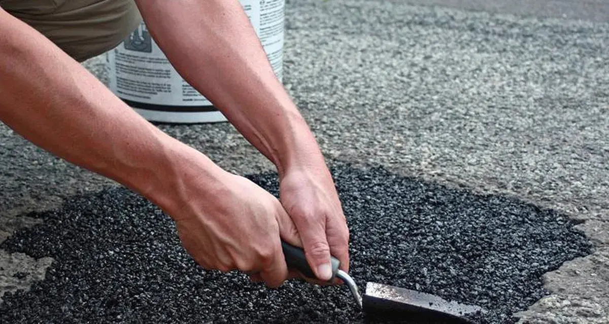 Händer som reparerar asfalt med en spade. En hink med "Asphalt" syns i bakgrunden.
