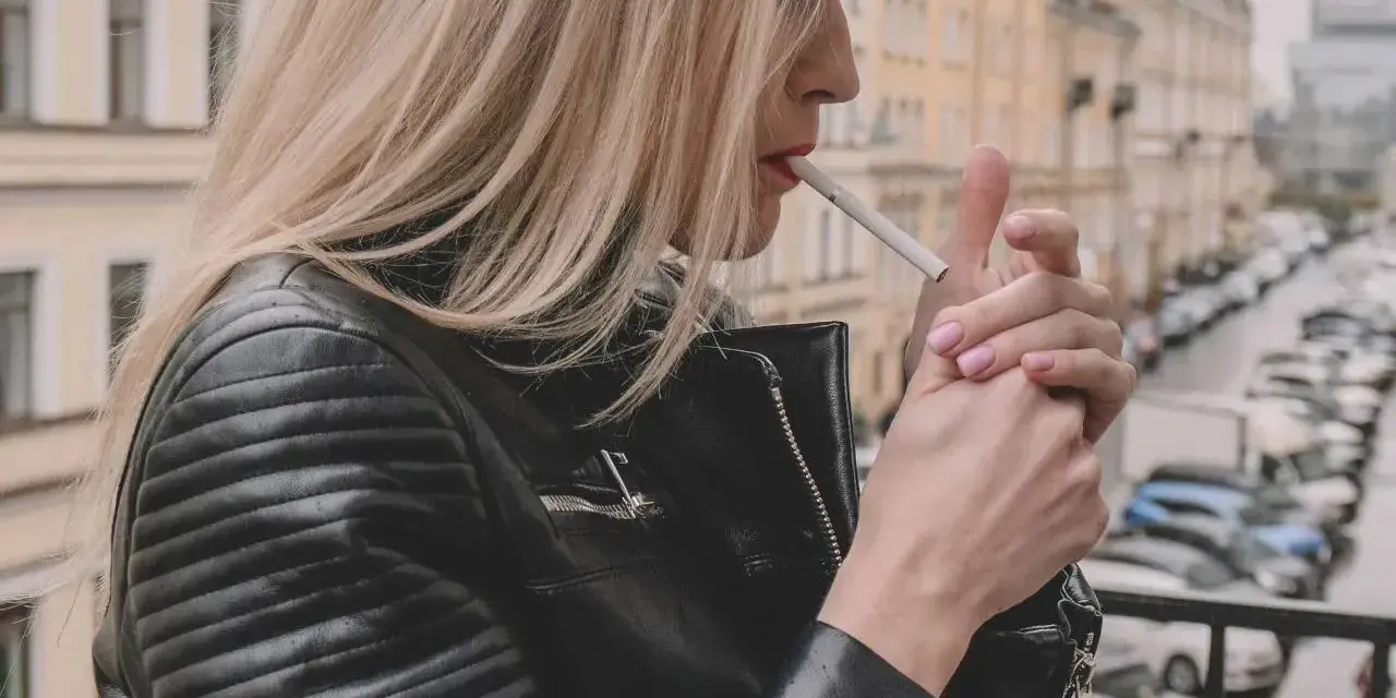 Kobieta w skórzanej kurtce zapala papierosa na balkonie, zastanawiając się, czy można palić na balkonie.