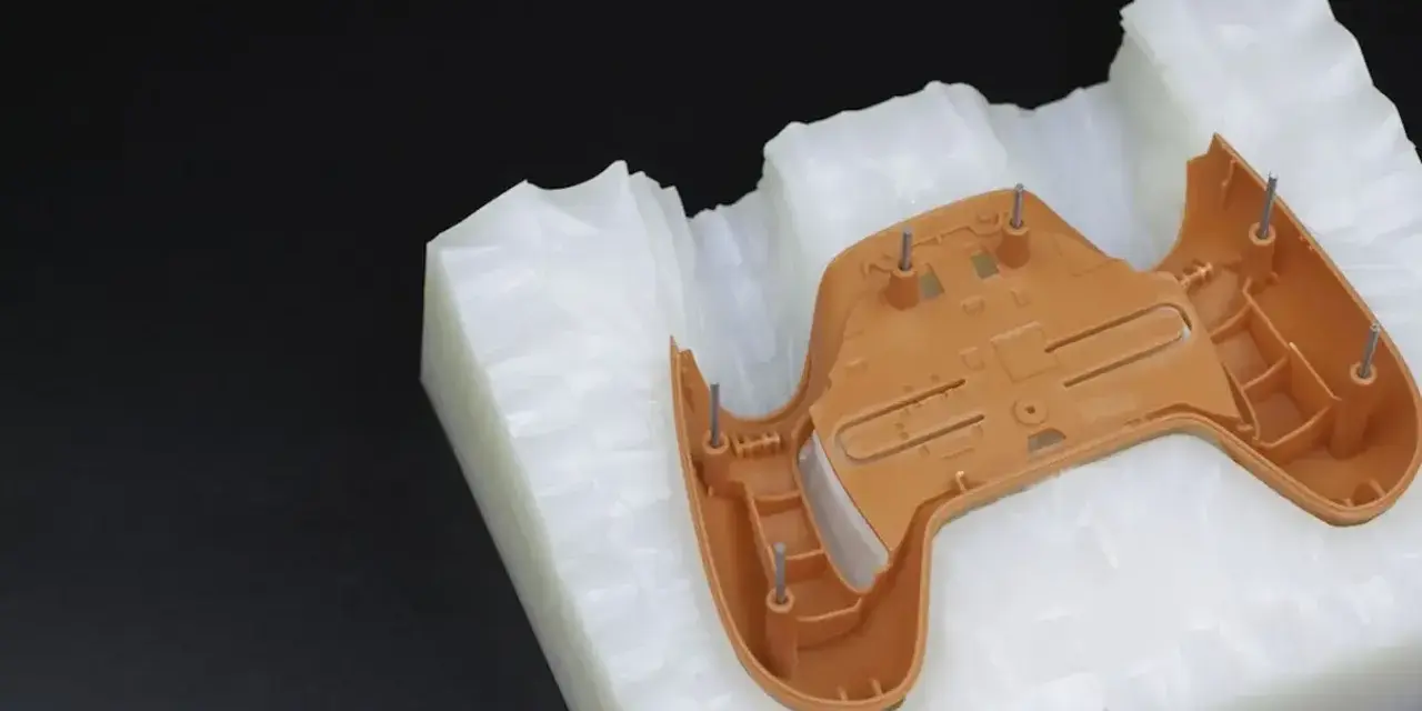 A brown plastic part, likely from a game controller, is placed on a white, textured material. This setup suggests a step in the polyurethane vacuum casting process, where a mold is being prepared.