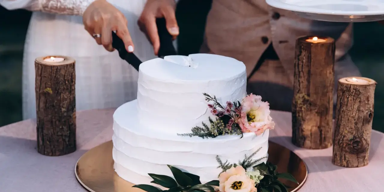 Biały tort weselny z teksturą, ozdobiony kwiatami. Para kroi tort, a obok płoną świece w drewnianych świecznikach. Trendy w prezentacji tortów weselnych to m.in. tekstury.