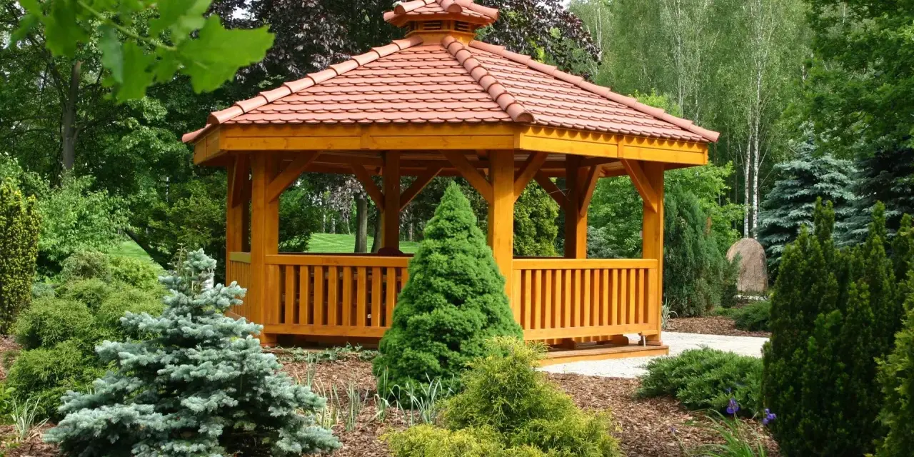 Drewniana altana z czerwonym, ceramicznym dachem, otoczona bujną zielenią. Idealne miejsce na relaks, a dach z dachówki to świetny pomysł, czym pokryć dach altany tanio i estetycznie.