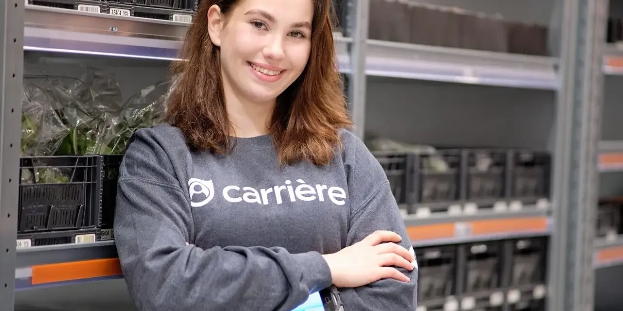 Young woman in a grey sweatshirt with "carrière" logo, holding a scanner, in a warehouse with shelves of black crates.